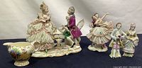 Five porcelain items arranged including a large Dresden figurine of a man and woman by a piano, two Victorian figurines, and a small floral cream pitcher