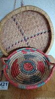 Photo showing top view of two woven baskets stacked, one coil round basket with colors and handles, one larger flat basket with woven straw pattern and colored accents.