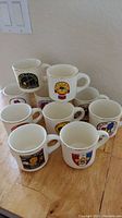 Lot of 10 white ceramic coffee mugs with Boy Scouts of America regional emblems printed on them, arranged in a stacked formation on a wooden floor.