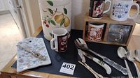 Photo showing large ceramic canister with fruit designs, assorted coffee cups, cutlery including a decorative spreader, and a floral patterned cheese board.