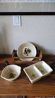 Three ceramic Poppytrail by Metlox serving pieces and wood bread server by Woodpecker Woodwear shown on wooden surface with wall light switch and framed art above.