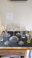 Photo showing eight assorted specialty glasses including Martini Rossi Millennium champagne flutes, margarita glasses with etched polka dot design, and smaller Bacardi Limon and Crown Royal glasses.