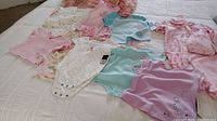 Overview of nine baby girl/toddler outfits laid out on a bed, mostly pastel colors such as pink, purple, blue, and white, showing variety of bodysuits and sleepers with hangers included