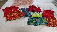 Lot of toddler boys clothing laid out on a white bedspread, consisting of red tops and red patterned shorts, blue Baby Gap jeans, green shirt, and blue patterned shorts, with hangers.