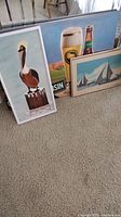 Three framed wall art pieces leaning against a wall: pelican painting, Wisconsin beer poster, and sailboat painting.
