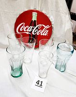 Photo showing the 12-inch red Coca-Cola metal wall decor in bottle cap design along with six Coca-Cola branded glasses of varying tints and materials