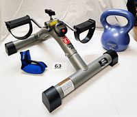 Stamina InStride Cycle XL mini exercise bike with black pedals and digital monitor, blue 10lb kettlebell, and blue/black wrist weight visible