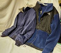 Two men's jackets, one navy LL Bean and one navy and black North Face, displayed hanging side by side showing full length