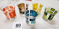 Five heavyweight mid-century cocktail glasses with vibrant enameled Rx patterns in orange, brown, yellow, blue, and green.