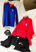 Four clothing items displayed together including blue and red NBA pullover jacket, red Antigua fleece pullover jacket, black short sleeve shirt with NBA Finals 2001 logo, and black NBA Philadelphia 76ers hat.