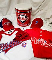 Photo showing two Phillies shirts, two baseball-style hats, and a metal Phillies logo can with white lid displayed together on a white cloth background.