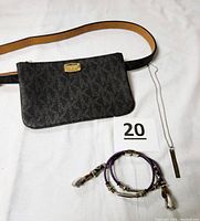 Front view of black Michael Kors monogram waist bag with attached tan adjustable belt, purple leather braided watchband, and silver chain necklace with pendant.
