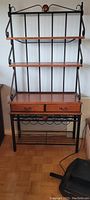 Front view of the baker's rack showing three wooden shelves, black wrought iron frame with decorative scrollwork, and wood accents.