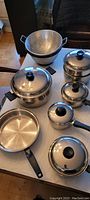 Full view of the stainless steel pots, pans, and colanders arranged on a table showing the entire cookware set.