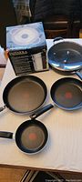 Photo of Lagostina Pastaiola pasta pot in box alongside three T-fal frying pans of different sizes and Starfrit frying pan with lid on a table.