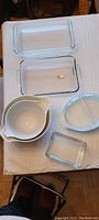 Nine Pyrex items displayed on white cloth, mix of clear glass bakeware, blue and white divided serving dish, and white nested mixing bowls.
