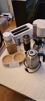 Photo showing small kitchen appliances on a table including toaster, kettle, coffee grinder, coffee maker, and wooden egg holder.