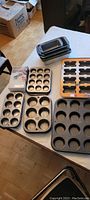 Photo showing 10 muffin tins and 6 loaf pans arranged on a table, including a new mini loaf pan in packaging and a rolling circle cutting set.