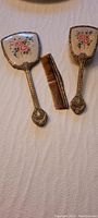 Vintage filigree brass hand mirror, hairbrush and comb laid out flat on a white cloth surface. Mirror and brush backs have petite point floral embroidery under glass.