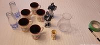 Overview of all glassware and collectibles including six mugs, two milk jugs, two Dutch figurines, etched vase, and teddy egg cup