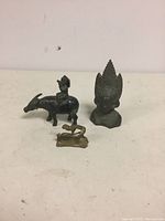 Three metal figures including boy on ox, decorative bust, and small animal figure arranged on tabletop.
