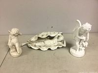 Three white bisque porcelain figures displayed: two cupids and the reclining nude within a shell for size and condition examination.