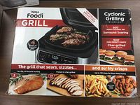 Box front showing Ninja Foodi Grill branding, grill with steaks, key selling points like cyclonic grilling technology and rapid air surround searing at 500°F