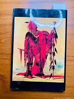 Front cover of the book featuring a full-length colored painting of a Native American man in traditional attire with red body paint and feathers, standing with cultural items.