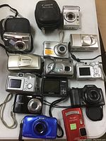 Collection of 14 various branded compact digital cameras laid out on table, showing multiple colors and styles including Canon, Kodak, Olympus, Sony, Pentax models with attached wrist straps and some cases