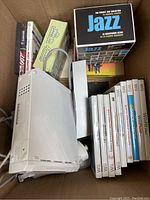 Box containing Nintendo Wii console with cables, multiple Wii game cases, boxed jazz CD collections, and Beatles CD set