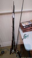Photo of multiple fishing rods leaning against a wall, one reel visible on table.