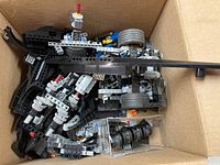 Box containing assorted Lego Technic pieces including large rubber wheels and gear components, showing mechanical structures and connectors.