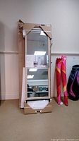 Full view of easel floor mirror in original packaging standing upright with beige carpet and wall background. Box shows some wear at corners.