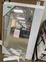 Mirror packaged in white foam corner protectors, wrapped in plastic, showing the brass-colored frame and reflective surface.