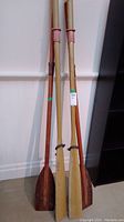 Full view of two pairs of wooden oars leaning against wall, showing length and style differences