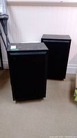 Pair of black rectangular speakers standing on carpet against wall