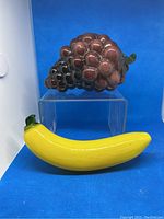 Photo of purple glass grape cluster positioned above yellow glass banana figurine on blue background