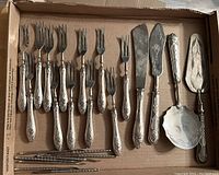 Full view of the assortment of stainless steel serving utensils laid out in a cardboard box including forks, knives, and servers