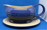 Blue ceramic Le Creuset gravy boat on matching cream-colored plate, side view showing 'Le Creuset' embossed logo and handle.