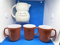 White ceramic hot chocolate pitcher labeled 'Chocolat Chaud' alongside three burnt orange Pyrex mugs.