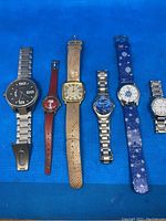 Photo of six wrist watches laid out horizontally on a blue surface, showing different styles and straps