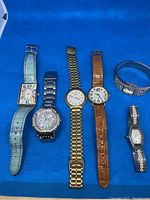 All six wrist watches arranged side by side on a blue surface, showing straps and faces