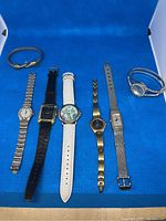 Lot contents showing six wrist watches arranged in a row with a gold tone elephant bangle on the left side, on a blue background.