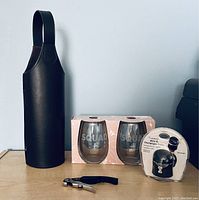 Front view of lot showing the brown pleather wine bottle holder, two boxed stemless glasses labeled SQUAD, corkscrew tool, and wine and champagne bottle sealer in package.