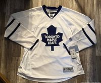 Front view of white Toronto Maple Leafs hockey jersey with Toronto Maple Leafs logo and blue trim on sleeves and collar.