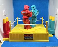 Photo showing the Rock’Em Sock’Em Robots toy with red and blue robots in a yellow boxing ring with corner posts in red, yellow, and blue.