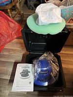 Black hard case opened, showing Simoniz polisher wrapped in clear plastic and green foam pad on lid, along with instruction manual on floor beside it.