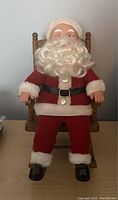 Santa Claus figure wearing red suit and black belt, seated in wooden rocking chair, on flat surface.