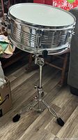 Full view of Dixon Percussion chrome snare drum on tripod stand with rubber feet, showing the drum's side and tension rods.