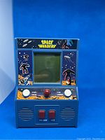 Front view of blue handheld Space Invaders game showing screen, control buttons and outer space-themed graphics